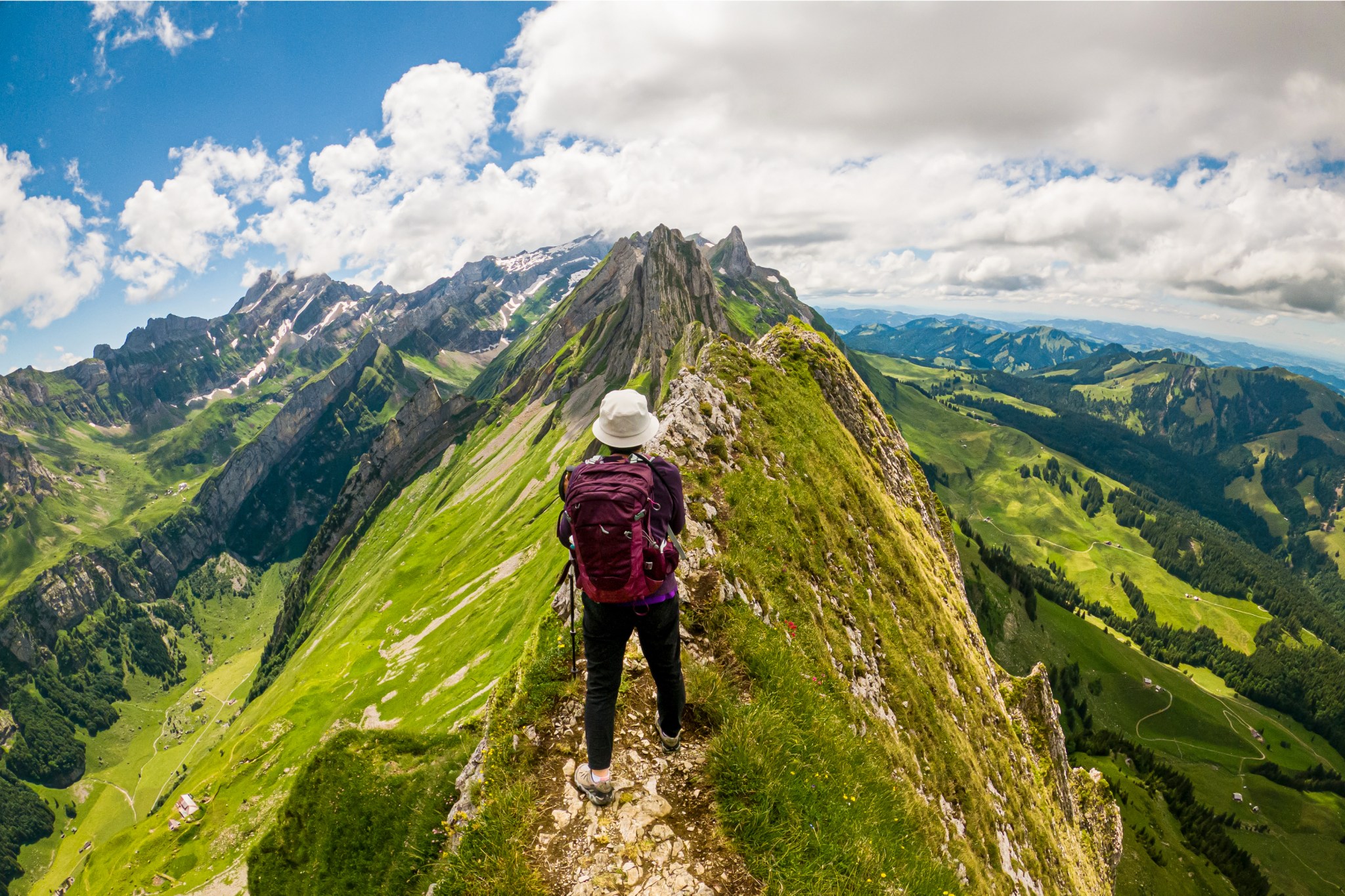 A solo hiker stands at the summit of a green mountain.