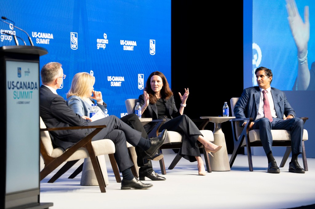 Four panelists on stage at the RBC US-Canada Summit. A person with dark hair dressed in all black speaks with both hands raised expressively in the center, while three other panelists — including one in a navy suit and red tie on the far right — listen. The branded US-Canada Summit backdrop and podium are visible in the background.