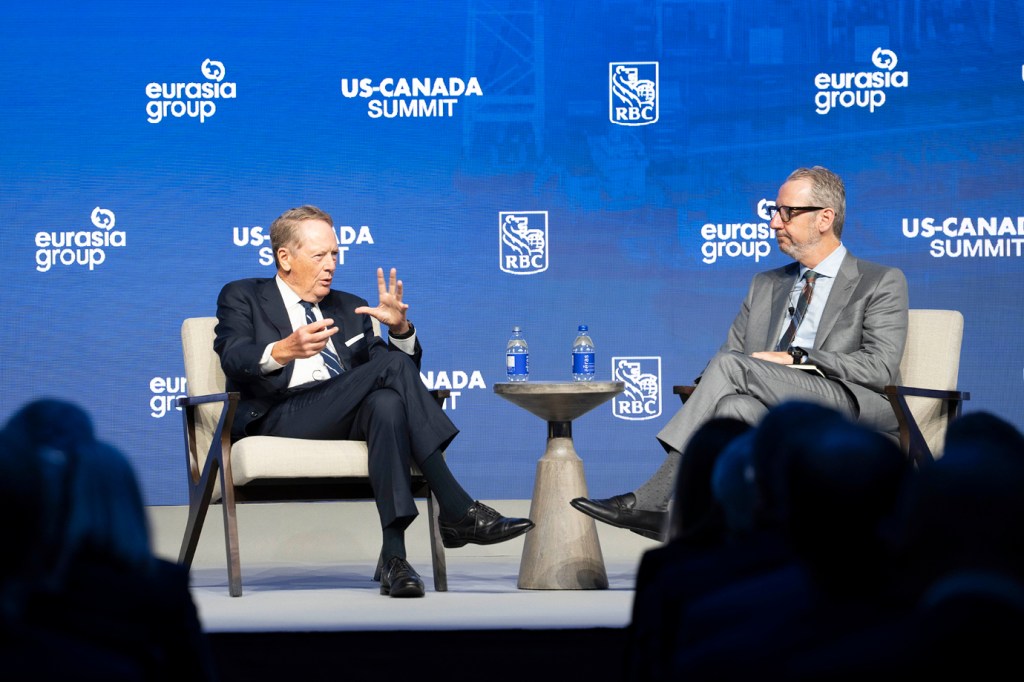 Two speakers in a fireside chat on stage at the RBC US-Canada Summit. An older person in a dark navy suit with a striped tie gestures with a raised hand while speaking on the left, while a person in a grey suit and glasses listens attentively on the right. A small round table with water bottles sits between them. Audience silhouettes appear in the foreground.