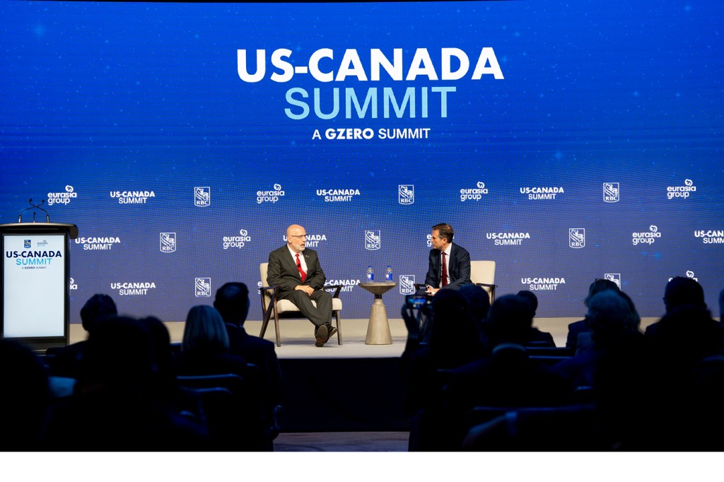 Wide-angle view of the main stage at the RBC US-Canada Summit, subtitled "A GZERO Summit." Two speakers are seated in chairs at center stage — one in a dark suit with a red tie and one in a dark suit — with the large illuminated summit backdrop dominating the background. A branded podium stands to the left. Audience members fill the foreground in silhouette.