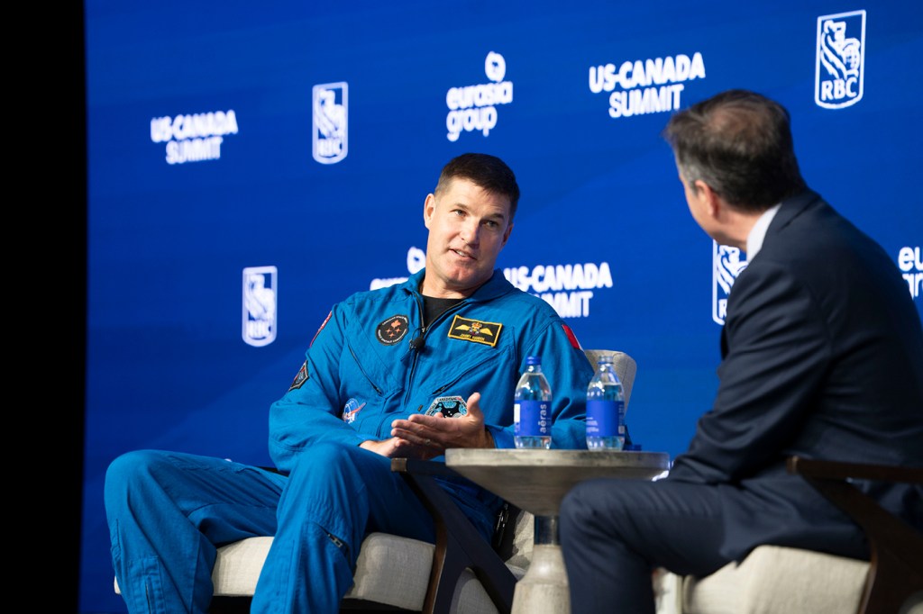 Close-up of two speakers in conversation on stage at the RBC US-Canada Summit. On the left, Canadian astronaut Jeremy Hansen is wearing a blue astronaut flight suit with mission patches speaks and gestures with their hands. A person in a dark suit, partially visible with their back to the camera, sits across from them on the right. Water bottles rest on a table between them against the blue branded backdrop.