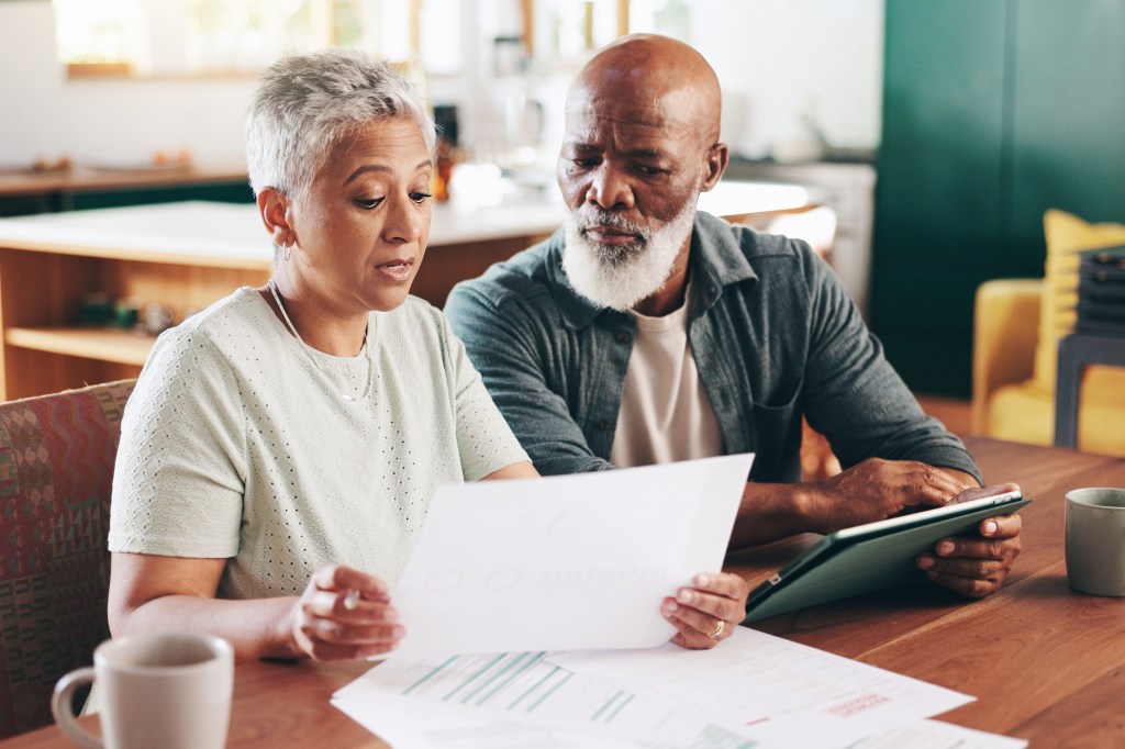 A mature couple is sitting down at a wooden table in the dining room looking at documents and a tablet. They have serious expressions.