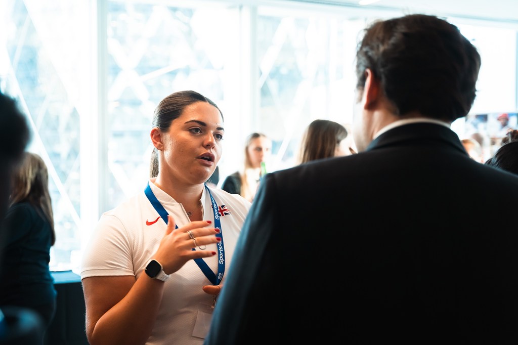 Katy Head is talking to a man in a room filled with people. Katy is a white woman, with long dark hair tied back into a ponytail. She is wearing a white short sleeve shirt, a white Apple watch and has a blue lanyard around her neck.