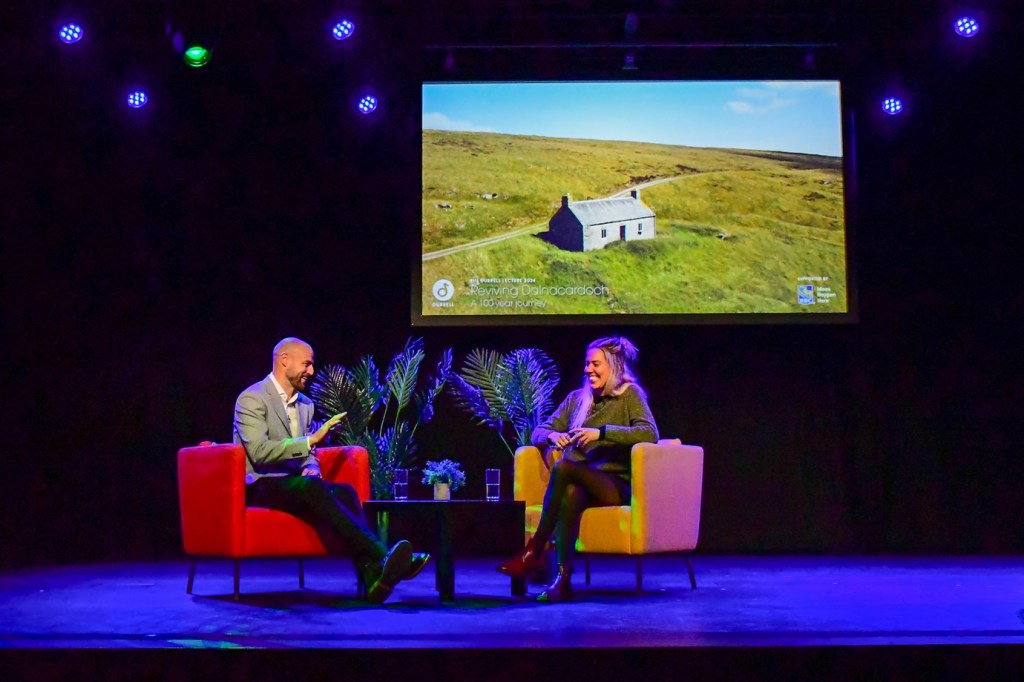 Two people are sitting on a stage in an auditorium, having a conversation. On one side is Mike Hudson, director of Conservation Knowledge at Durrell Wildlife Conservation Trust. Mike is a white man. He is bald and has a brown beard. He is wearing a grey blazer over a white button-up shirt, and black pants. He is sitting on a red chair and talking to a woman, Libby Penman, a wildlife filmmaker. Libby is a white woman. She has long blond hair, and is wearing a grey sweater, rights and dark red boots. She is sitting on a yellow chair and is smiling. There is a black table between them with two glasses of water and a small plant sitting on the table. There are two large ferns sitting behind them. In the background is a screen showing a picture of part of the Dalnacardoch estate. The image shows a small house surrounded by green grass.