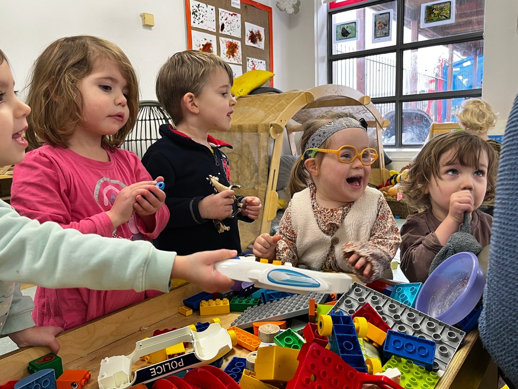 Five young children are playing with toys in a nursery centre. The children are standing in front of a wooden table that is filled with toys such as big Lego blocks. One of the kids is wearing yellow glasses and has a big smile on her face. Behind them is a wall covered in children's art, a wooden tent-like structure with pillows inside, and a box full of stuffy toys. A large window shows part of the outside with a play structure.