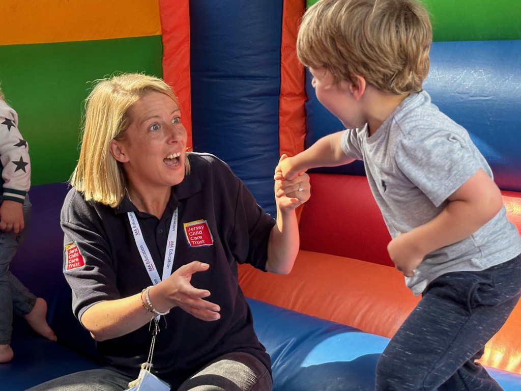 A woman, who is a support worker for Jersey Child Care Trust, holds the hand of a young boy at a local pre-school. The two of them are in a colourful bouncy castle. The woman has shoulder-length blond hair and is wearing a dark shirt with the JCCT logo, grey jeans, a couple of bracelets and a white lanyard around her neck. She is looking at the boy with her mouth open. The boy has short blond hair and is wearing a grey t-shirt and dark grey sweatpants. He is holding on to the woman's hand.