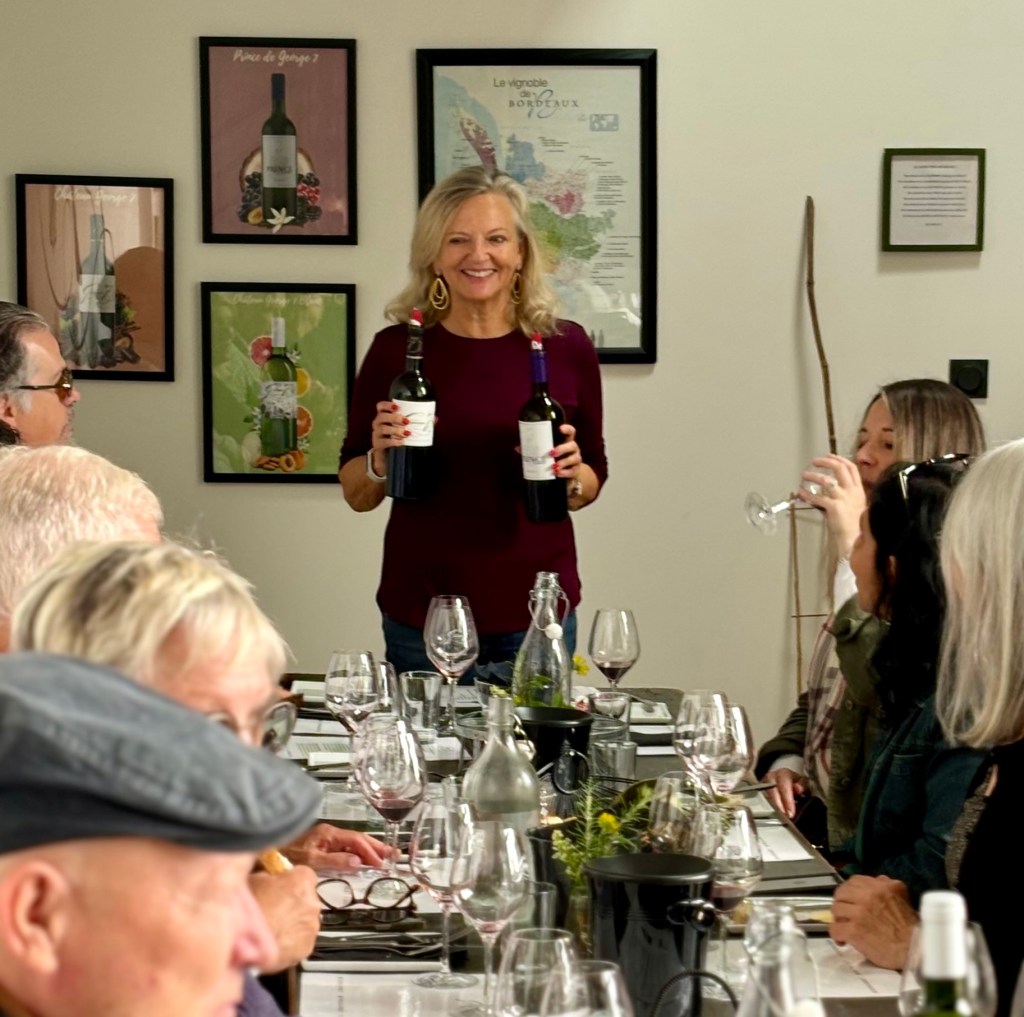 A woman with blonde hair, wearing a burgundy long-sleeve top and gold earrings, stands at the head of a long dining table holding a red wine bottle in each hand and smiling at the camera. Seated guests on both sides of the table have wine glasses, water carafes, and tasting materials in front of them. One guest to the right is sipping from a glass. The wall behind the host features several framed wine-related art prints and a large map labeled "Le vignoble de Bordeaux."