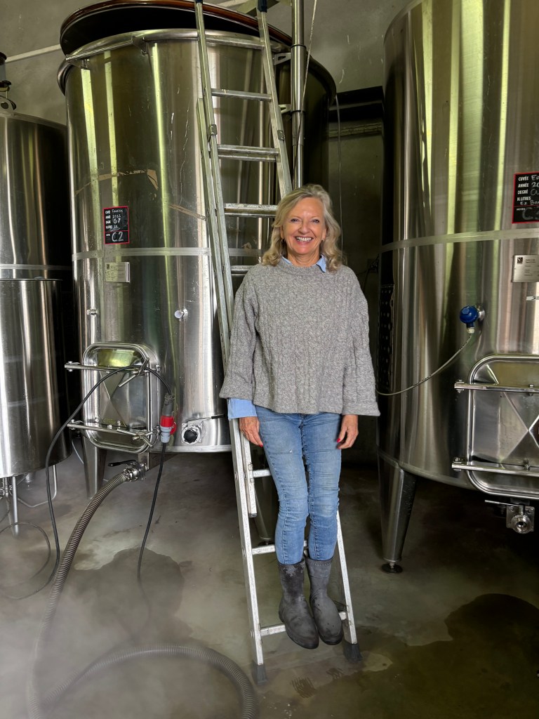 A smiling person stands on the bottom rung of a metal ladder between large stainless steel fermentation tanks in a winery cellar. They are wearing a gray cable-knit sweater over a blue collared shirt, jeans, and rubber boots. Labels with production details are visible on the tanks in the background.