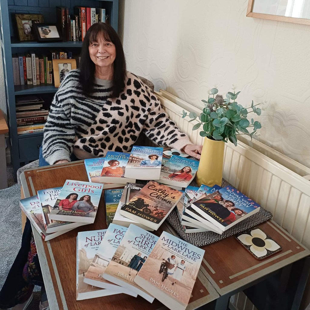 A smiling woman with long dark hair, wearing a leopard-print sweater, sits at a wooden table covered with approximately 15 paperback novels, all authored by Pam Howes. Visible titles include The Midwives of Lark Lane, The Shop Girls of Lark Lane, Cathy's Clown, The Liverpool Girls, A Royal Visit to Victory Street, and several books in the Mersey Square series. A blue bookshelf filled with books stands behind her, and a yellow vase with eucalyptus sprigs sits to her right.