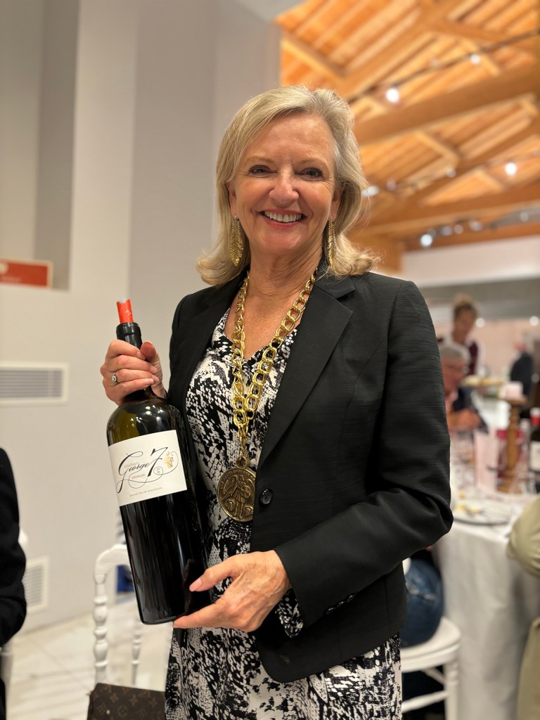 A smiling woman with shoulder-length blonde hair stands in a bright event venue, holding a large bottle of Château George 7 Bordeaux red wine. She is dressed in a black blazer over a black-and-white patterned dress and wears a chunky gold chain necklace with a large medallion. The background shows a warmly lit room with wooden ceiling beams and guests seated at round tables.