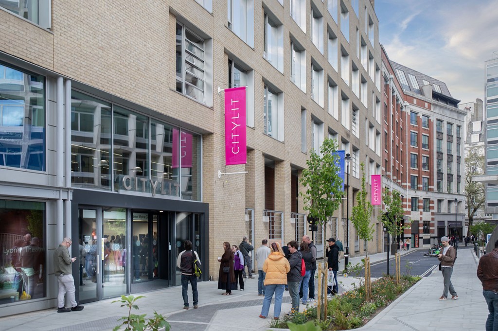 The exterior of City Lit college in London, featuring a modern multi-storey building with light brick cladding and large glass windows. Pink vertical banners displaying the "CITYLIT" name hang along the façade. A group of people stand and mingle on the pedestrianised pavement outside the entrance, with young trees lining the street. Additional buildings and a blue sky with light clouds are visible in the background.