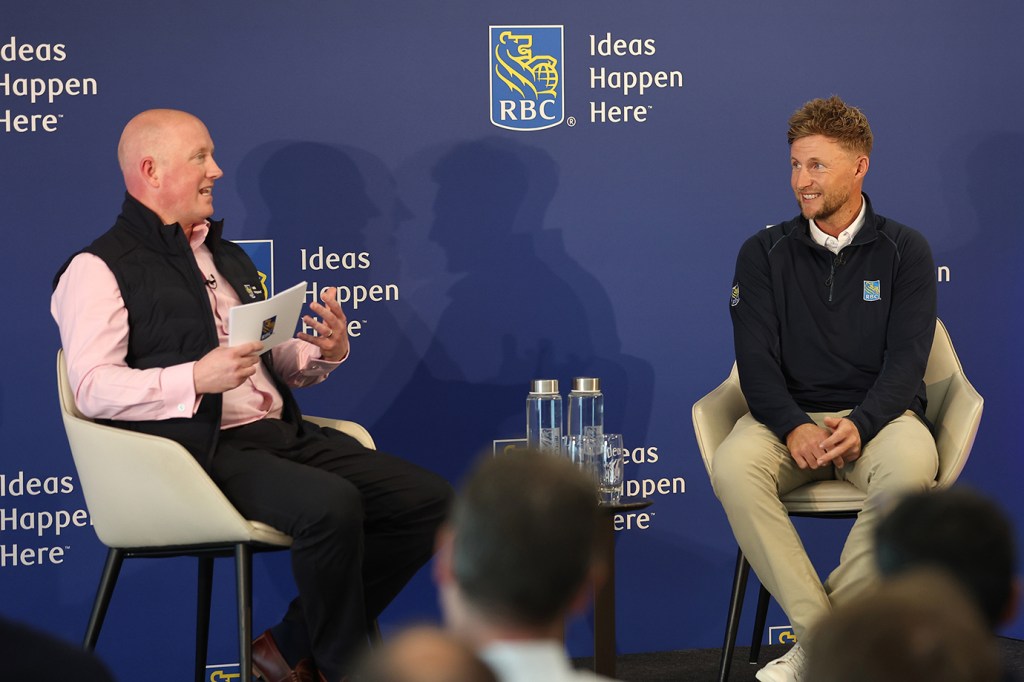 Two people in an on-stage interview, seated in cream armchairs facing each other against a deep blue RBC "Ideas Happen Here" backdrop, with water bottles on a side table between them and audience silhouettes in the foreground.