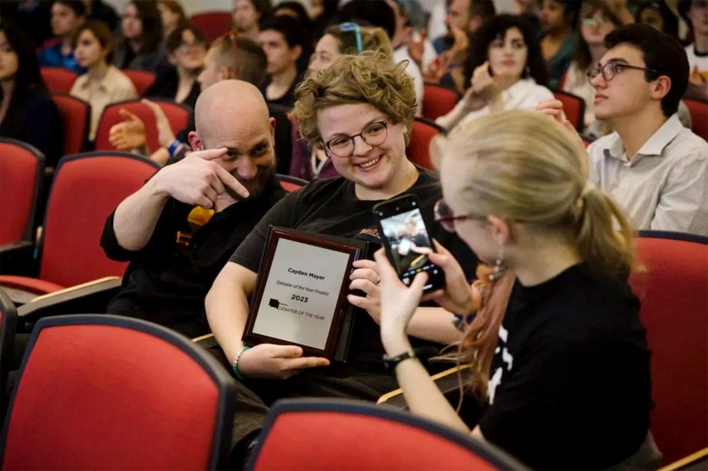 Three people are sitting in red chairs in a theater. The person in the middle is a white person with short, curly blond hair and glasses named Cayden Mayer, who is holding a plaque that says "Debater of the Year Finalist." Mayer is posing for a photo being taken by a blond woman on her left using a smart phone. The person on Mayer's right, a white man, is also posing for the photo. He is smiling and pointing a finger down. Mayer is smiling for the photo. There are about a dozen people sitting in the chairs behind them.