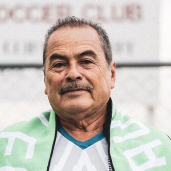 A headshot of Ismael Guzmán, a middle-aged man with short greying dark hair, brown eyes and a moustache. He is staring right at the camera with a serious expression. He is wearing a white and grey jersey with a light green and white scarf around his shoulders.