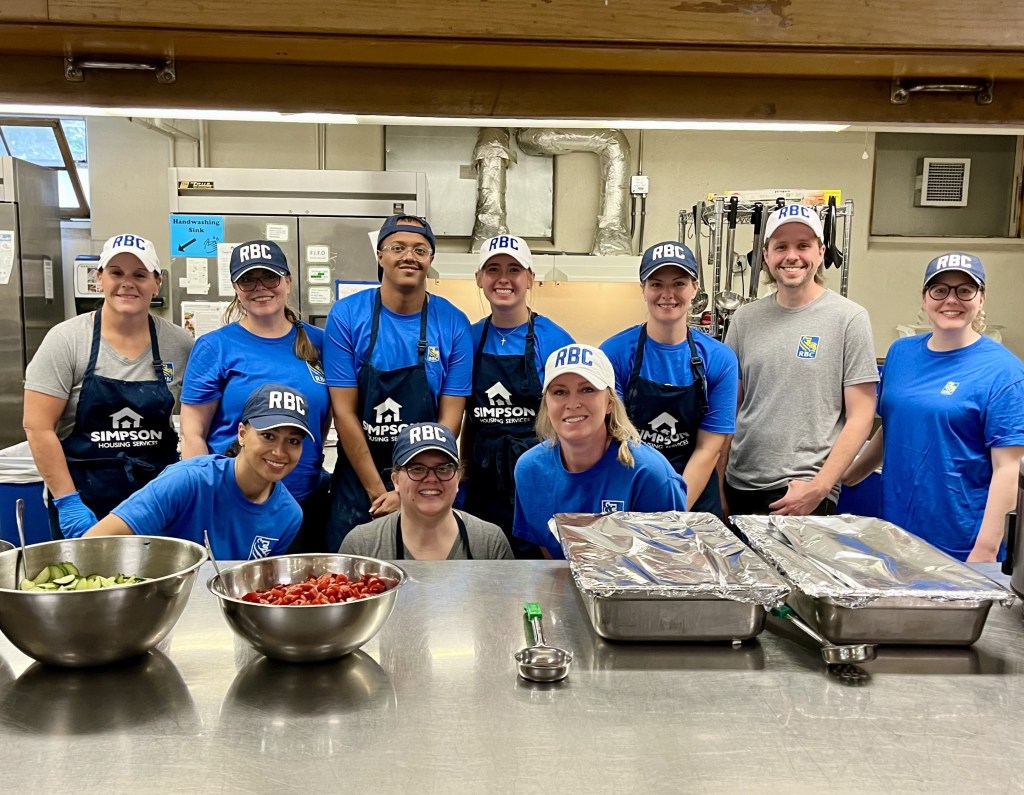 10 RBC employees are standing side-by-side, smiling at the camera. They are inside a large kitchen at Simpson Housing Services in Minneapolis. They are standing in front of a prep table, which has two silver bowls of cut-up vegetables and two large platters of covered food. Behind them are a large silver double-door fridge and various kitchen-wear.
