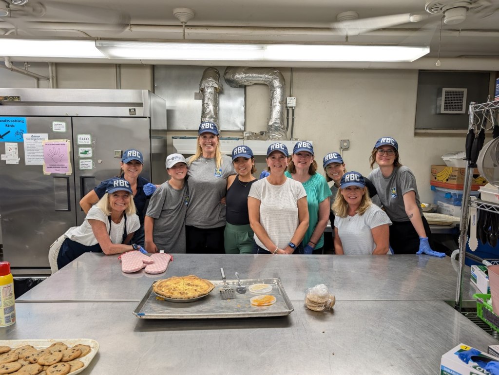 Nine RBC employees, and one youth, are standing side-by-side, smiling at the camera. They are inside a large kitchen at Simpson Housing Services in Minneapolis. They are standing in front of a prep table, which has oven mitts and a pie on it. Behind them are a large silver double-door fridge and various kitchen-wear.
