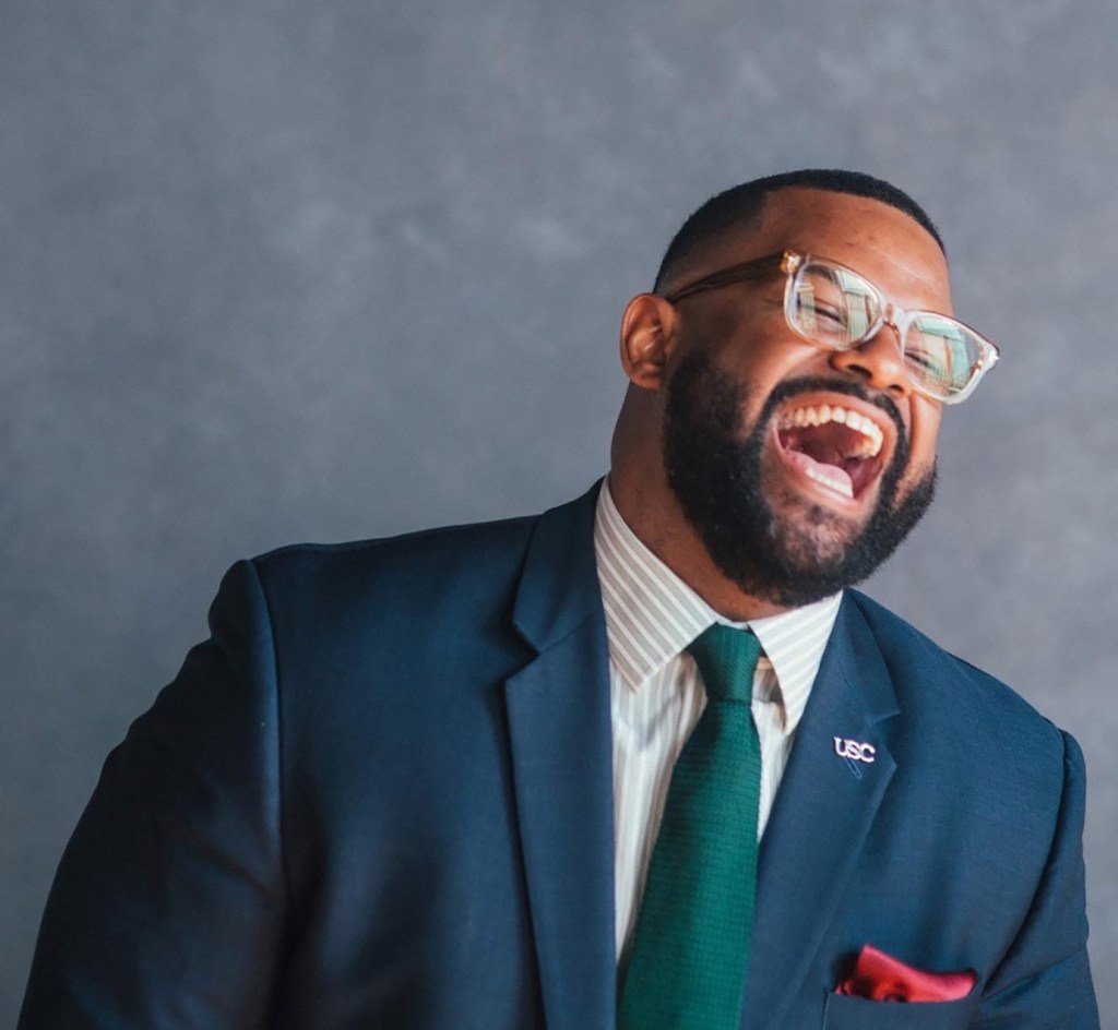 A professional portrait of a Black man with a short beard, wearing a navy blue suit jacket, a green knit tie, a white striped dress shirt, and a red pocket square. He has clear-framed glasses and a USC lapel pin, and is laughing with his head tilted slightly upward against a soft gray studio background.