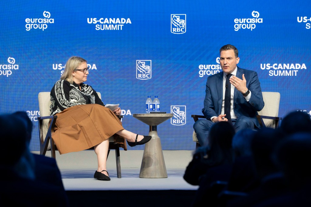 Deux conférenciers assis sur des chaises sur scène lors d’une discussion au coin du feu au Sommet RBC États-Unis-Canada.   Une personne aux cheveux blonds et aux lunettes portant un haut à motifs noirs et une jupe brune tient des notes à gauche, tandis qu’une personne en costume bleu foncé gesticule tout en parlant à droite.   La toile de fond de marque bleue affiche les logos du Sommet États-Unis-Canada, rbc et Eurasia Group.   Les silhouettes du public apparaissent au premier plan.