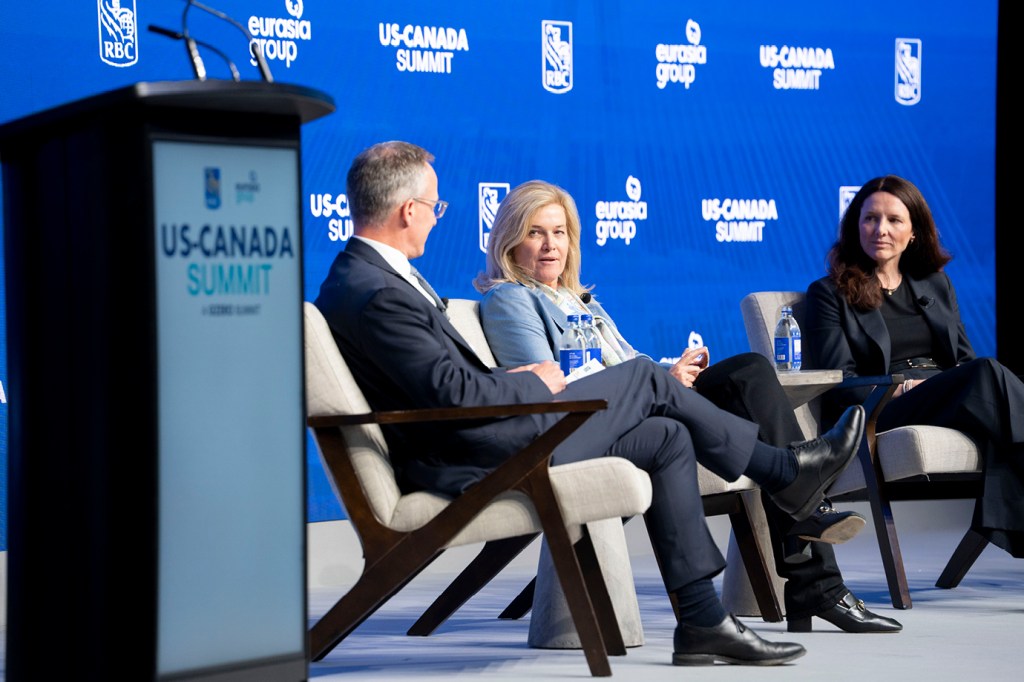 Trois panélistes assis sur des chaises sur scène au Sommet RBC États-Unis-Canada.   Une personne en costume sombre s’assoit le dos à la caméra à gauche, tandis qu’une personne aux cheveux blonds dans un blazer bleu clair parle au centre, et une personne aux cheveux foncés dans un blazer noir écoute à droite.   Un podium de marque du Sommet États-Unis-Canada est visible au premier plan.