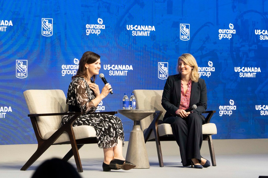 Deux femmes ont participé à une discussion au coin du feu sur scène au Sommet RBC États-Unis-Canada.   À gauche, une personne aux cheveux foncés dans une robe à motifs noirs et blancs tient un microphone et parle avec un sourire.   Sur la droite, une personne aux cheveux blonds dans un blazer noir et un chemisier floral tient un microphone et sourit en écoutant.   Une petite table ronde avec des bouteilles d’eau se trouve entre eux sur le fond de marque bleue.